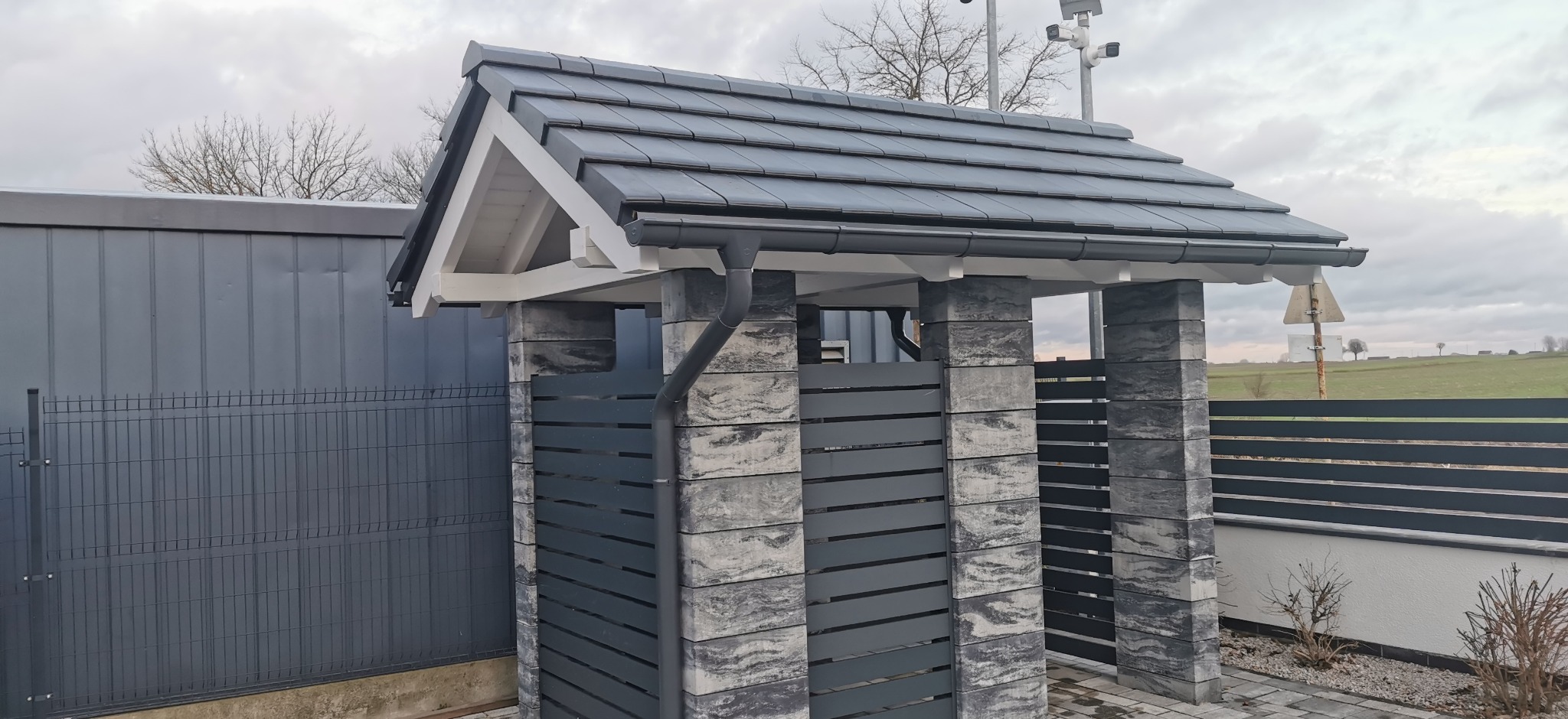 Zadaszenie nad bramą z szarej dachówki i rynnami, wsparte na kamiennych słupkach z poziomymi, szarymi panelami. W tle widoczne ogrodzenie panelowe i krajobraz.