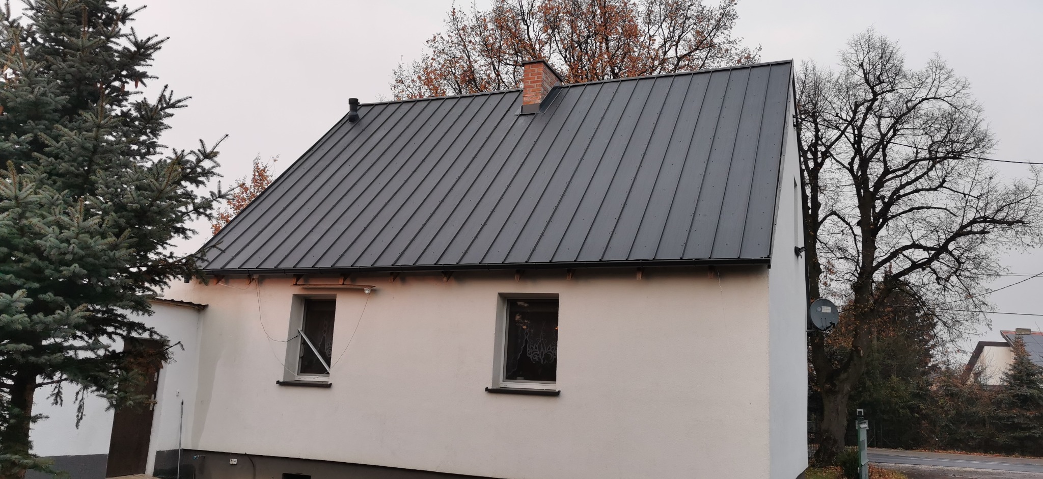 Dom z nowym, ciemnoszarym dachem z blachy na rąbek stojący. Widoczne drzewa bez liści i iglaste. Biała elewacja budynku. Ujęcie z poziomu gruntu.