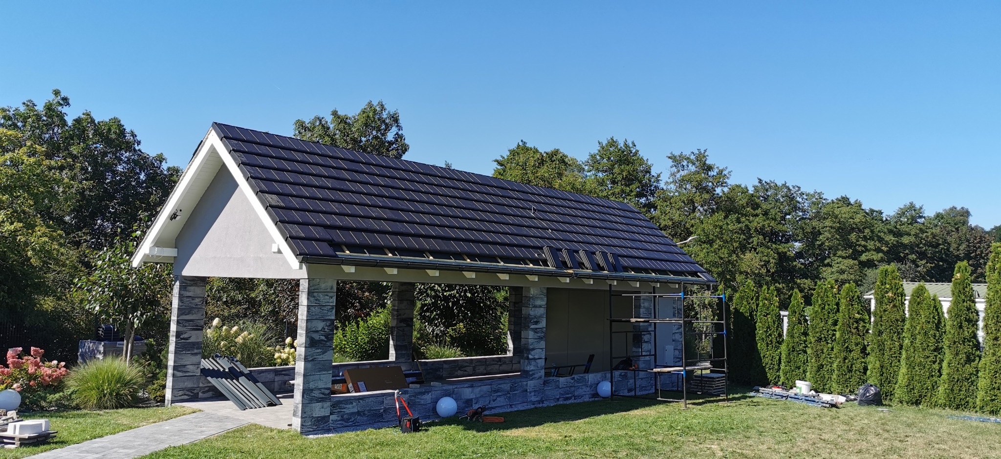 Ciemna dachówka ceramiczna na altanie z kamiennymi filarami, częściowo pokryta, z rusztowaniem i narzędziami na trawniku. Błękitne niebo w tle.