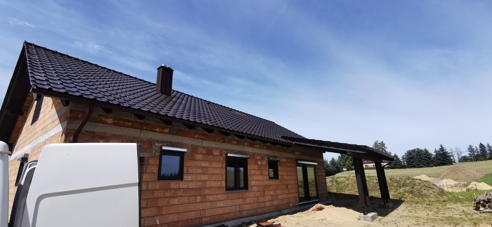 Dom w trakcie budowy z ciemnym dachem i rynnami. Widoczne cegły, okna z roletami i fragment białego samochodu dostawczego. W tle błękitne niebo.