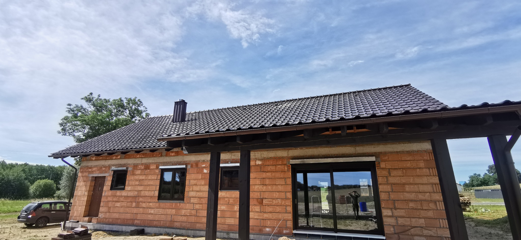 Dom w trakcie budowy z ciemną dachówką i orynnowaniem. Widoczny fragment elewacji z cegły, okna z roletami zewnętrznymi i fragment ogrodu w tle.