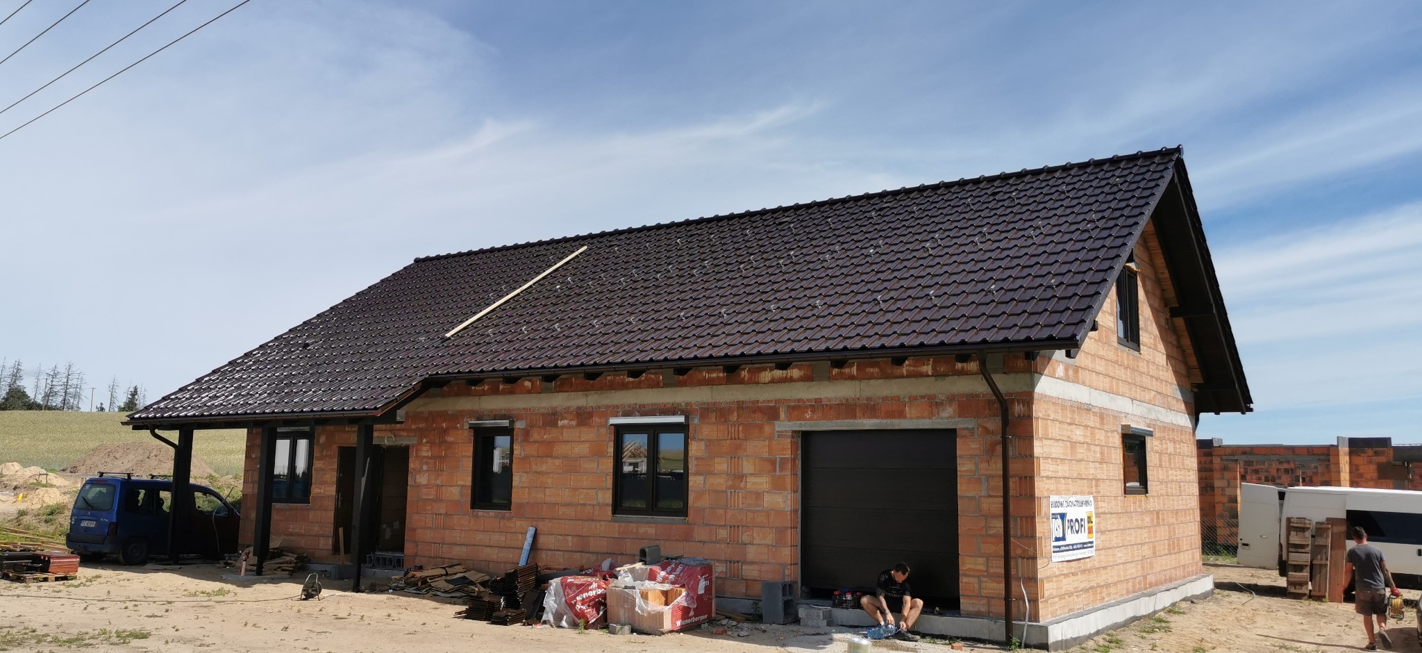 Dom w trakcie budowy z ciemnobrązowym dachem z dachówki ceramicznej. Widoczne rynny i fragment drewnianej łaty na dachu. Na pierwszym planie materiały budowlane.