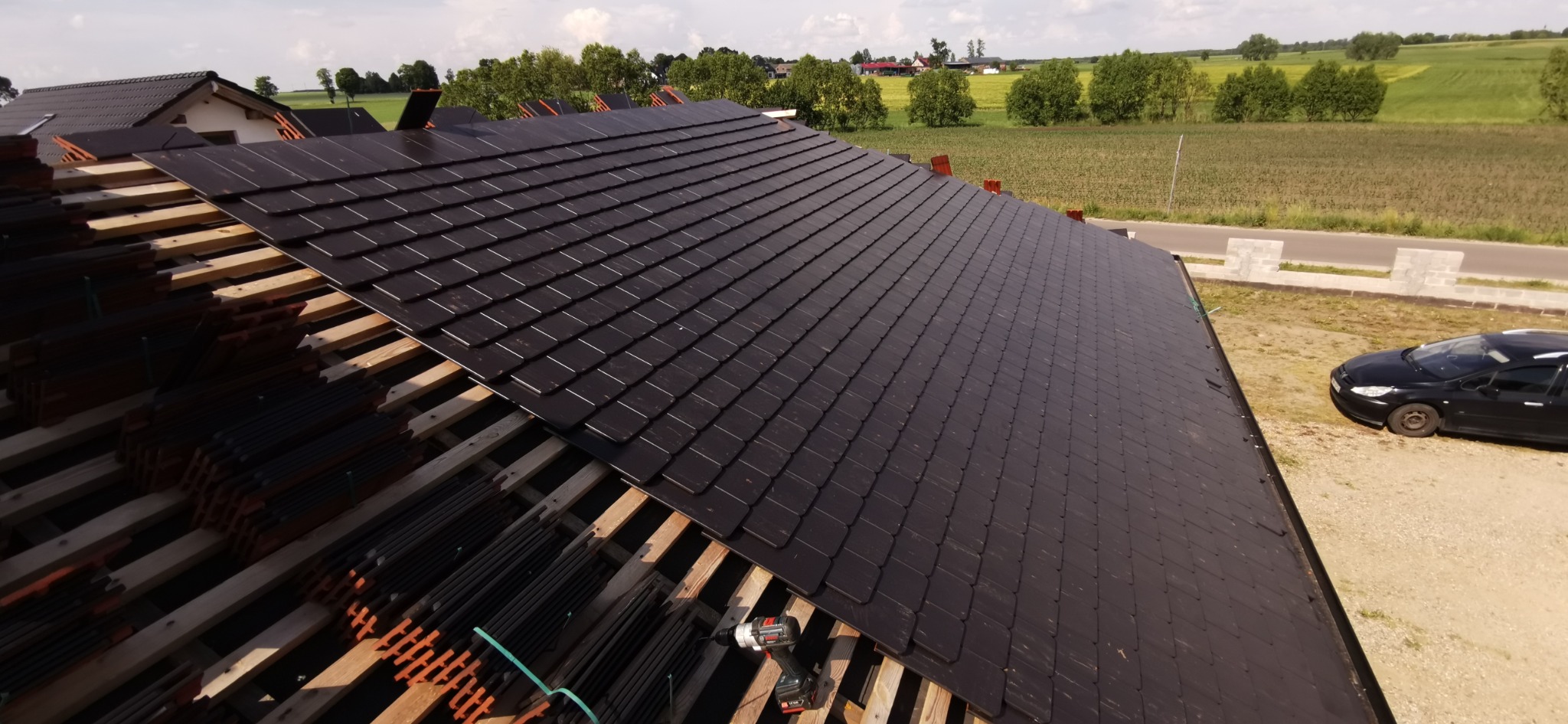 Układanie ciemnej dachówki na drewnianej konstrukcji dachu. Widoczne narzędzia pracy i częściowo ułożony dach na tle zieleni i błękitnego nieba.