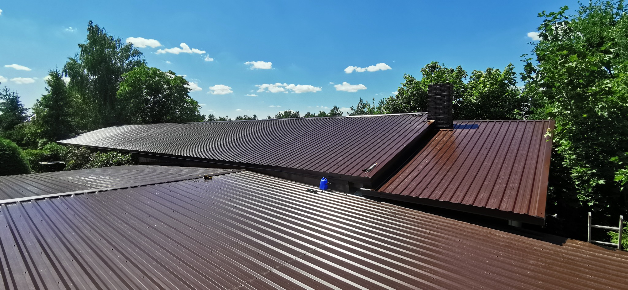 Brązowy dach z blachy trapezowej, widok z góry na tle błękitnego nieba i zieleni drzew. Na dachu widoczne narzędzia i pojemnik, sugerujące trwające prace.