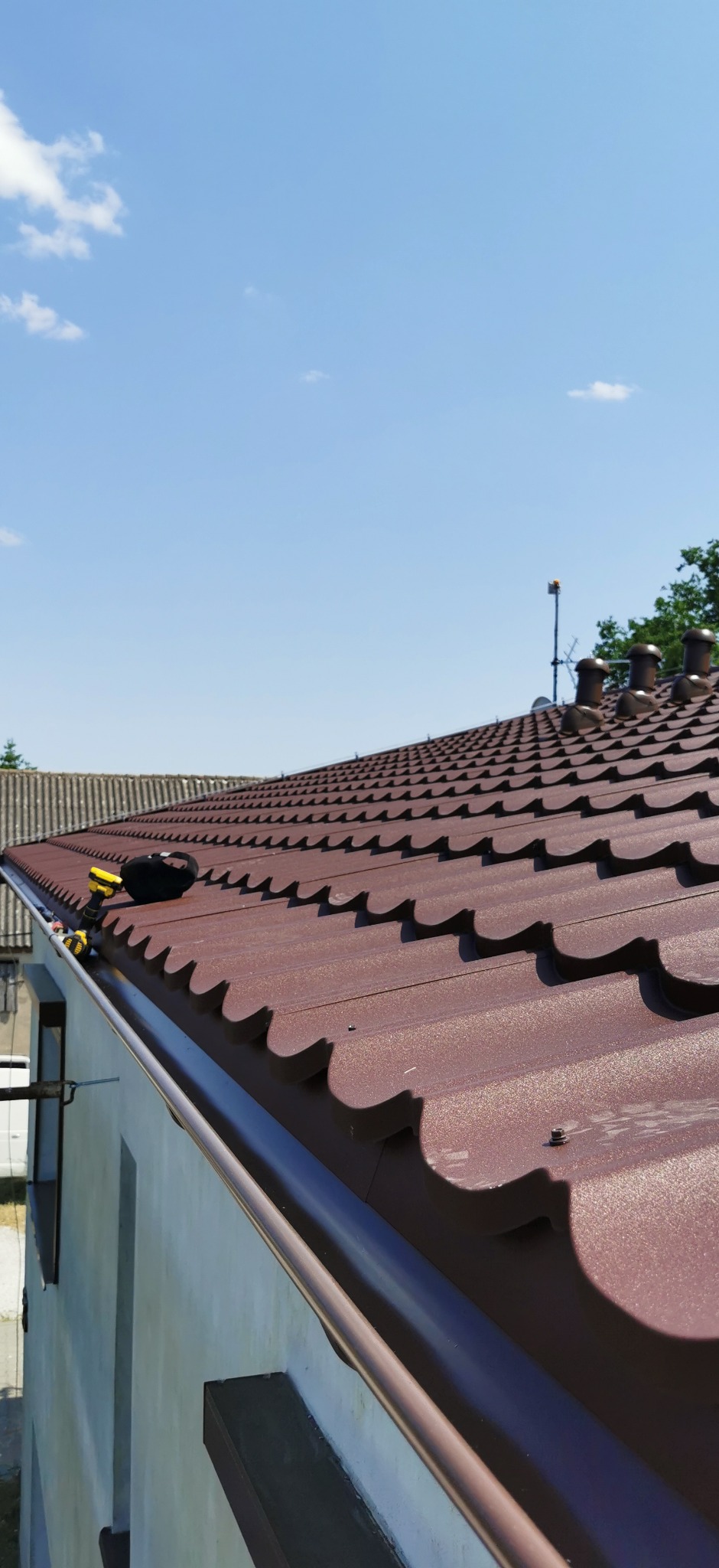 Ciemnobrązowy dach z blachodachówki modułowej, widok z dołu, z zamontowaną rynną w kolorze dachu. Na dachu leżą narzędzia dekarskie. Błękitne niebo w tle.