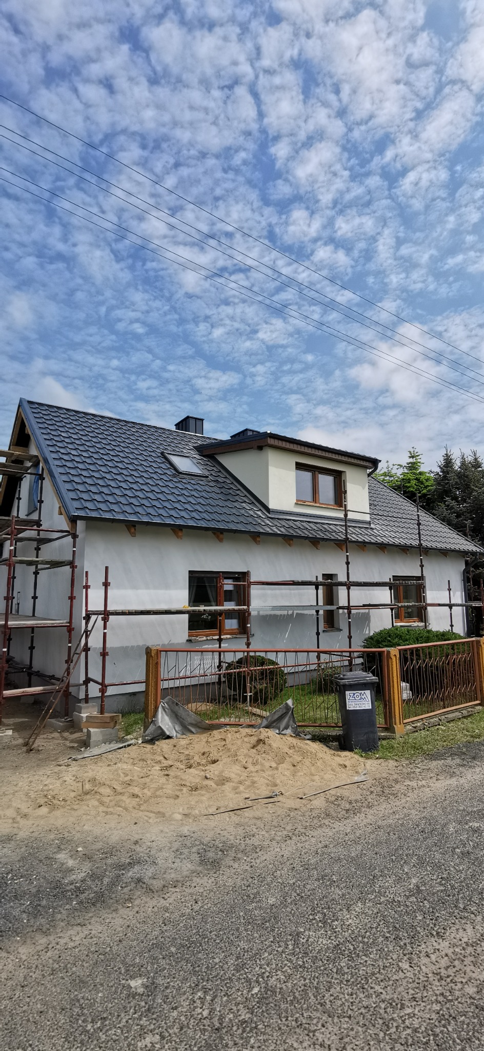Dom jednorodzinny z ciemnoszarą blachodachówką, częściowo otoczony rusztowaniem. Widoczny fragment ogrodzenia i sterta piasku. Błękitne niebo w tle.