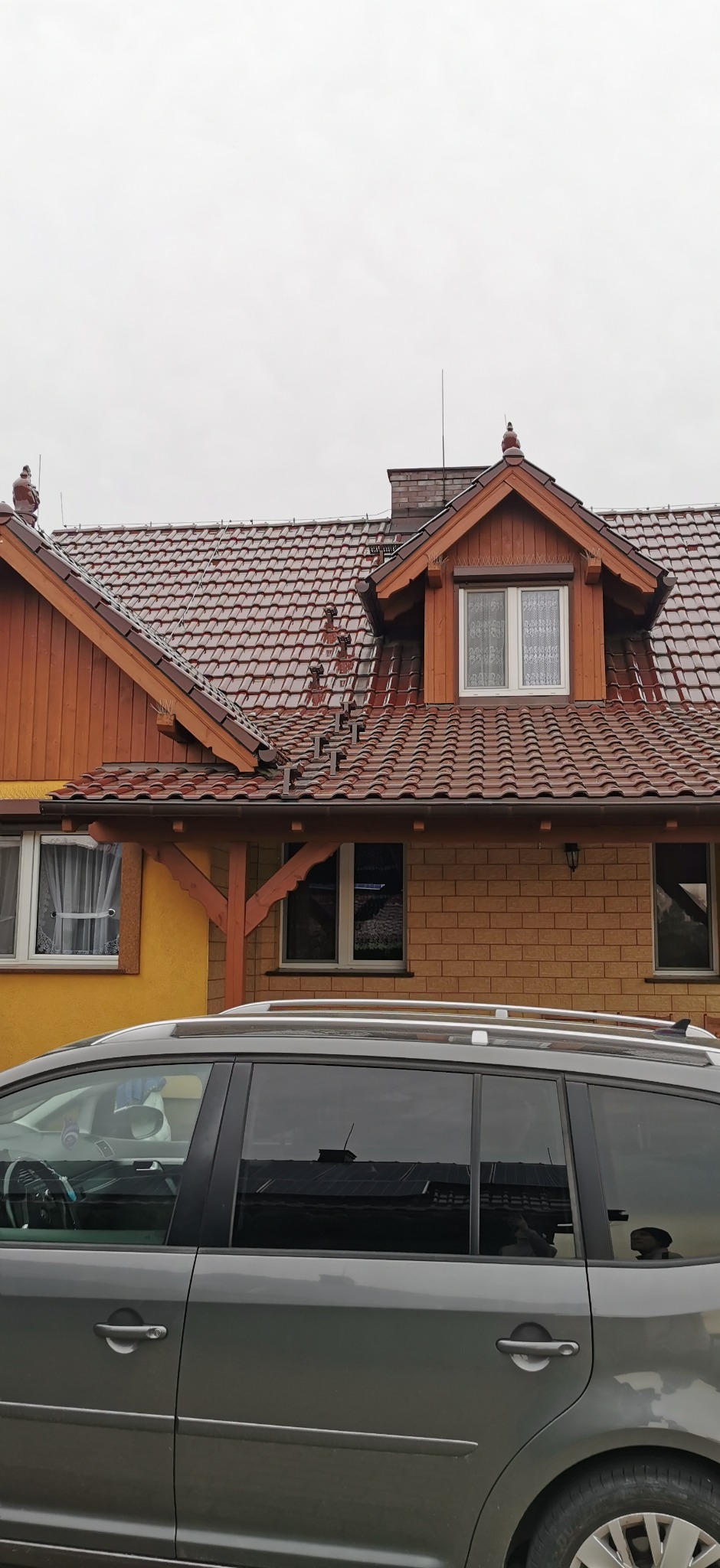 Dom z brązową dachówką ceramiczną, widoczne zabezpieczenia przeciw ptakom na dachu i rynnach, szara osobówka na pierwszym planie, pochmurne niebo w tle.