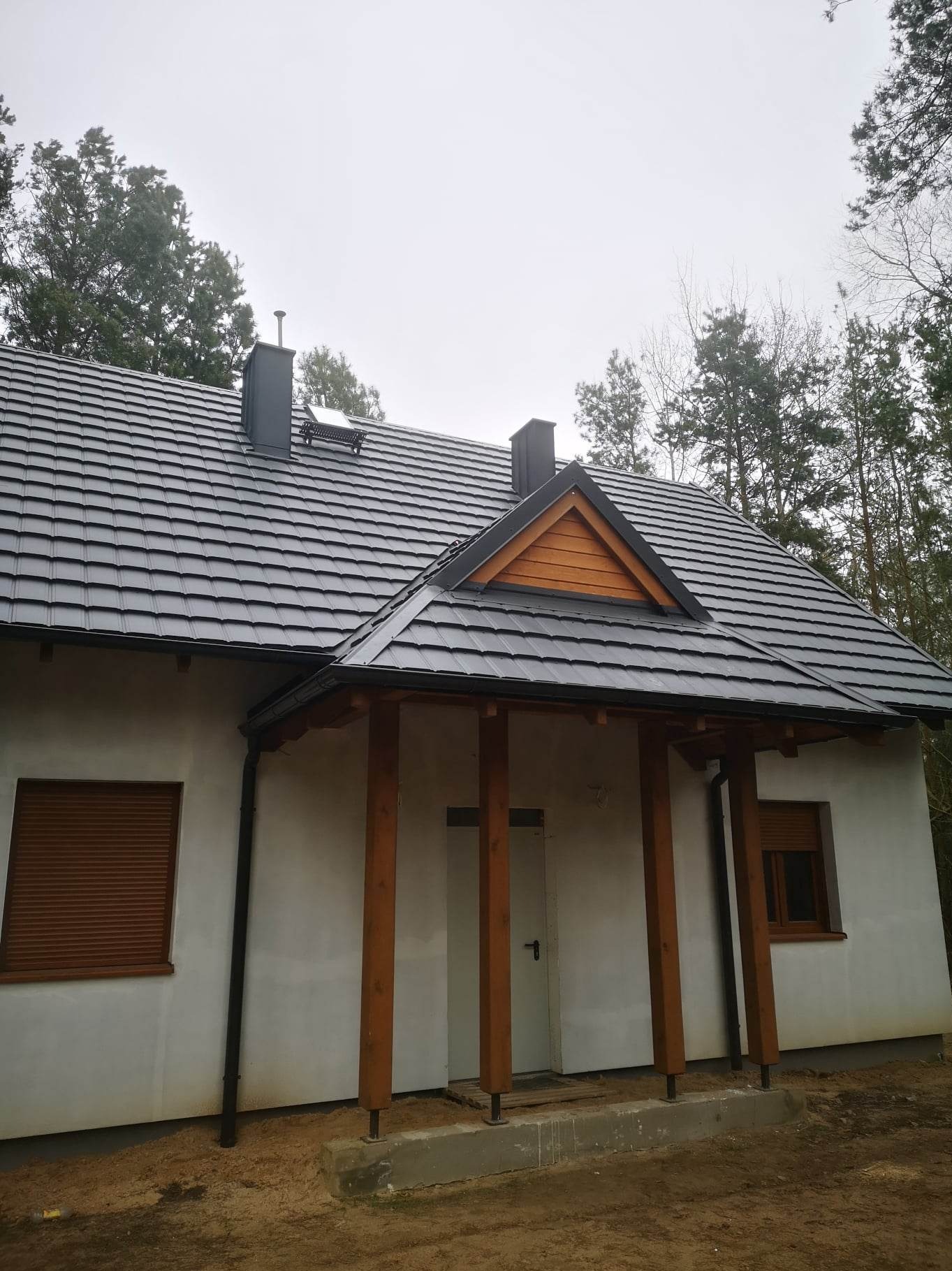 Dom jednorodzinny z nowym, grafitowym dachem z dachówki ceramicznej, drewnianą podbitką i czarnymi rynnami. Widoczne kominy i okno dachowe.