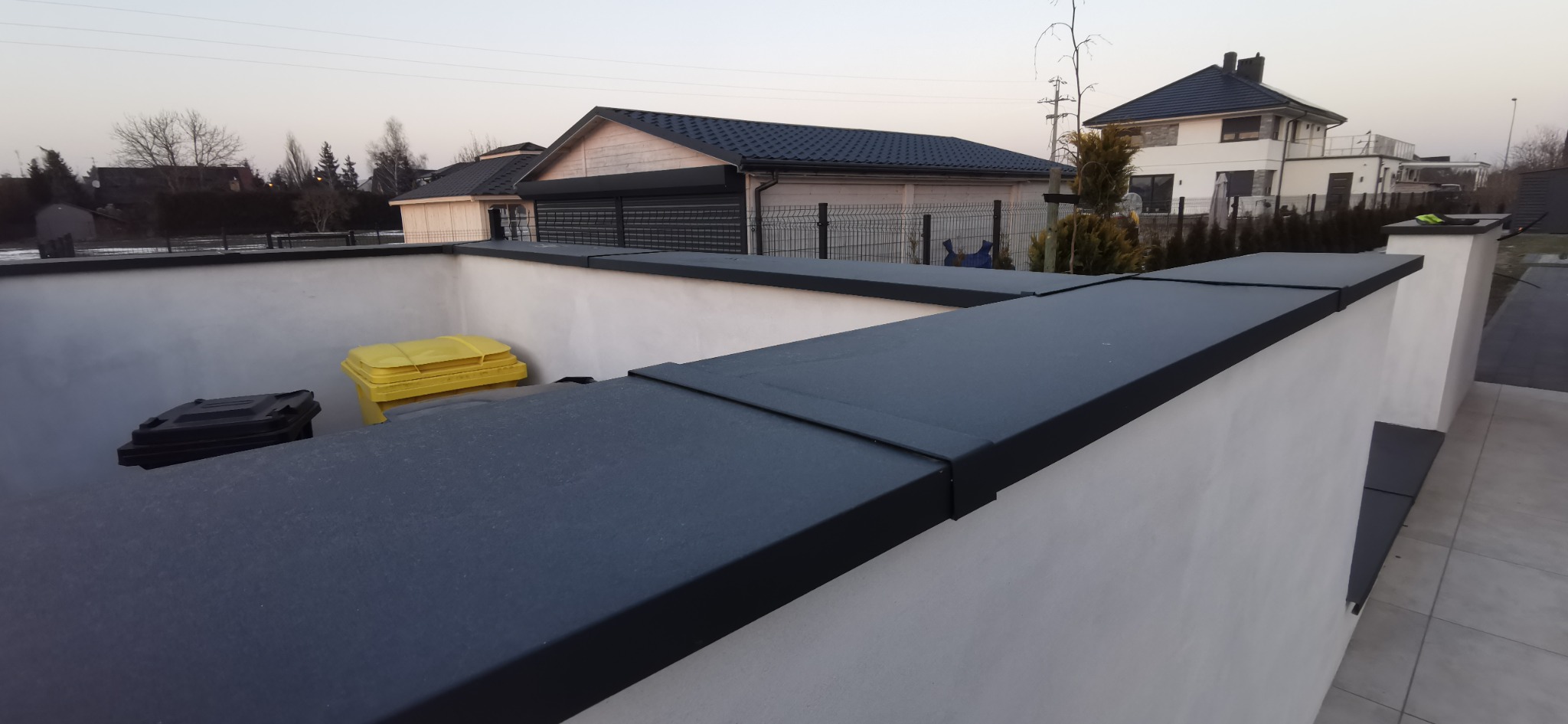 Obróbki blacharskie na murku oporowym z widocznymi pojemnikami na śmieci w kolorze czarnym i żółtym. W tle domy jednorodzinne i drzewa.