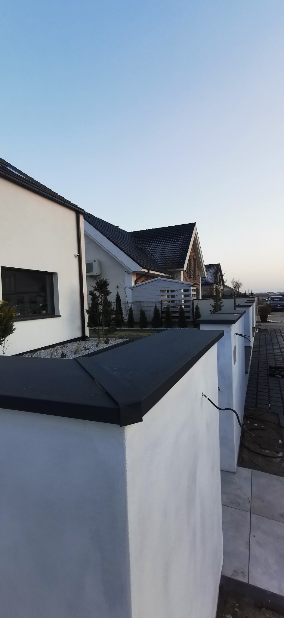 Nowoczesne domy jednorodzinne z ciemnymi dachami i białymi ścianami, widoczne z perspektywy ogrodzenia z czarnym wykończeniem. Minimalistyczny design i spokojna kolorystyka.