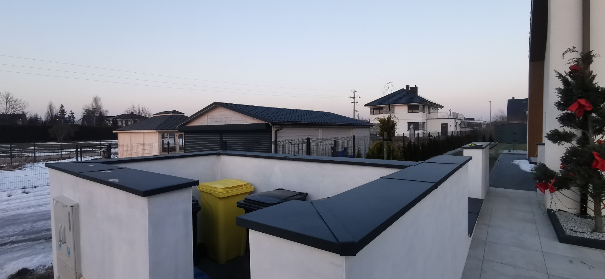 Nowoczesne obróbki blacharskie na murku okalającym śmietnik, z widokiem na domy z ciemnymi dachami w tle, w zimowej scenerii.