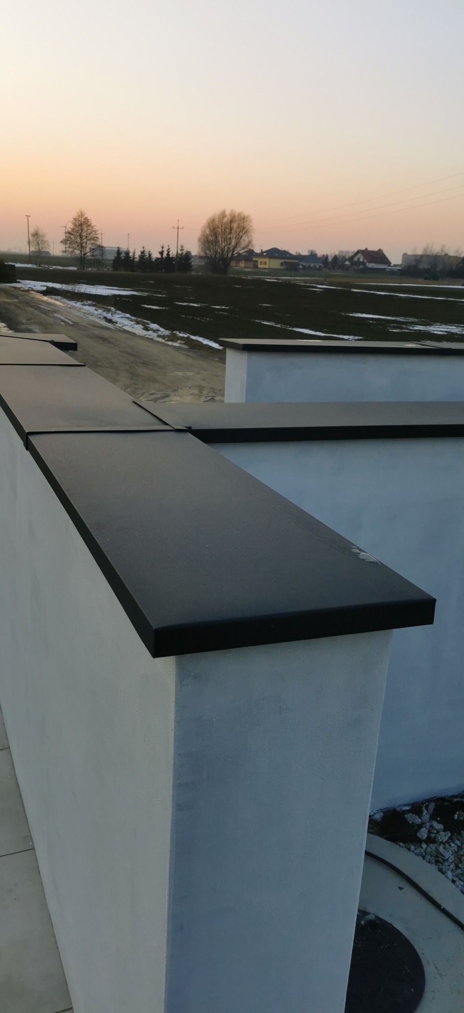 Nowoczesne obróbki blacharskie na murze oporowym w kolorze antracytowym, z widokiem na pole i zachodzące słońce. Estetyczne wykończenie.