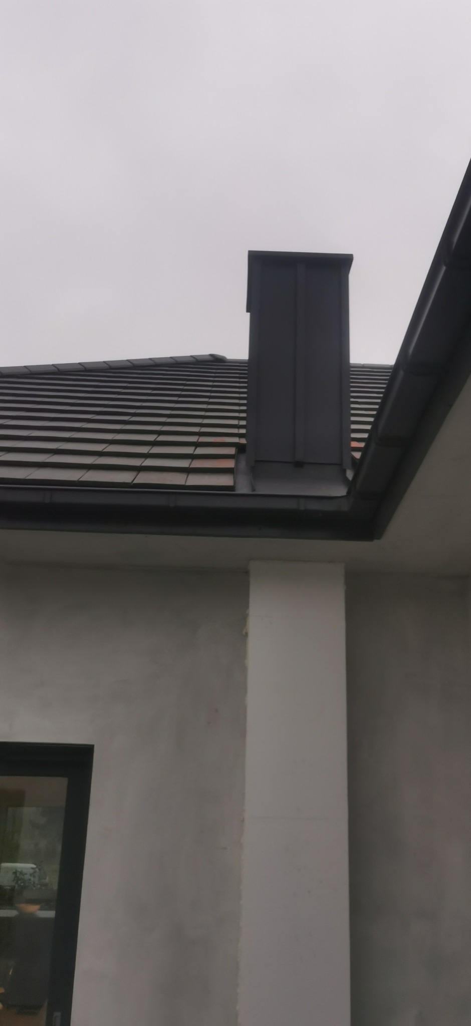 Szary dach z dachówki ceramicznej i komin z blachy na tle pochmurnego nieba. Widoczna ściana budynku z oknem i rynna.