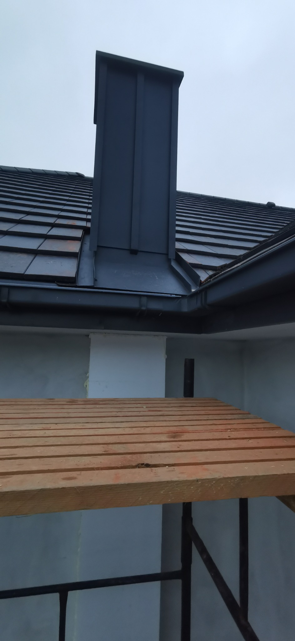 Nowy, grafitowy komin z blachy na tle dachu z dachówki ceramicznej i szarego nieba. Widoczny fragment rusztowania.