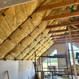 MIK-HAJ Usługi ogólnobudowlane Mikołaj Hajdacz - Skośny dach z drewnianą konstrukcją i żółtą izolacją, widok z rusztowania. Fragment otynkowanej ściany. Okno trójkątne w tle.