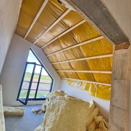 MIK-HAJ Usługi ogólnobudowlane Mikołaj Hajdacz - Poddasze w Pniewach w trakcie ocieplania. Widoczne belki, folia paroizolacyjna i wełna mineralna. Okno dachowe z widokiem na pole. Prace izolacyjne w toku.