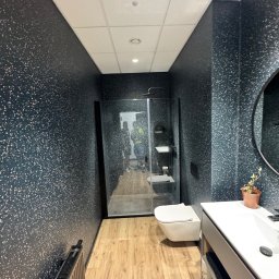 MIK-HAJ Usługi ogólnobudowlane Mikołaj Hajdacz - Nowoczesna łazienka z kabiną prysznicową typu walk-in, umywalką z szafką i toaletą. Ściany pokryte ciemną tapetą w jasne kropki, drewnopodobna podłoga. W tle widoczni pracownicy.