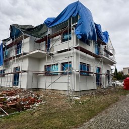 MIK-HAJ Usługi ogólnobudowlane Mikołaj Hajdacz - Biały dom w rusztowaniach, zabezpieczony plandekami, po świeżym malowaniu elewacji. Na pierwszym planie gruz i kostka brukowa, pochmurne niebo w tle.