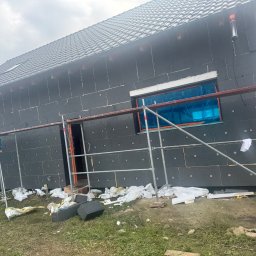 MIK-HAJ Usługi ogólnobudowlane Mikołaj Hajdacz - Dom w trakcie ocieplania elewacji czarnym styropianem, widoczne rusztowania i zabezpieczone okna folią. Na ziemi resztki materiałów budowlanych.