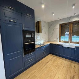 MIK-HAJ Usługi ogólnobudowlane Mikołaj Hajdacz - Granatowa zabudowa kuchenna z złotymi uchwytami, drewniany blat i podłoga imitująca drewno. Marmurowe płytki na ścianie i okno z drewnianymi roletami.