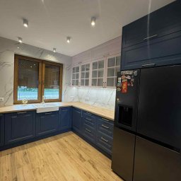 MIK-HAJ Usługi ogólnobudowlane Mikołaj Hajdacz - Elegancka kuchnia w odcieniach granatu i bieli z drewnianymi blatami. Widoczne szafki, okno z roletami i czarna lodówka z magnesami. Parkiet imitujący drewno.