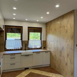 MIK-HAJ Usługi ogólnobudowlane Mikołaj Hajdacz - Jasna kuchnia z drewnianą ścianą i oknami z koronkowymi firankami. Uwagę przykuwają brązowo-beżowe płytki podłogowe i minimalistyczne, czarne uchwyty szafek.