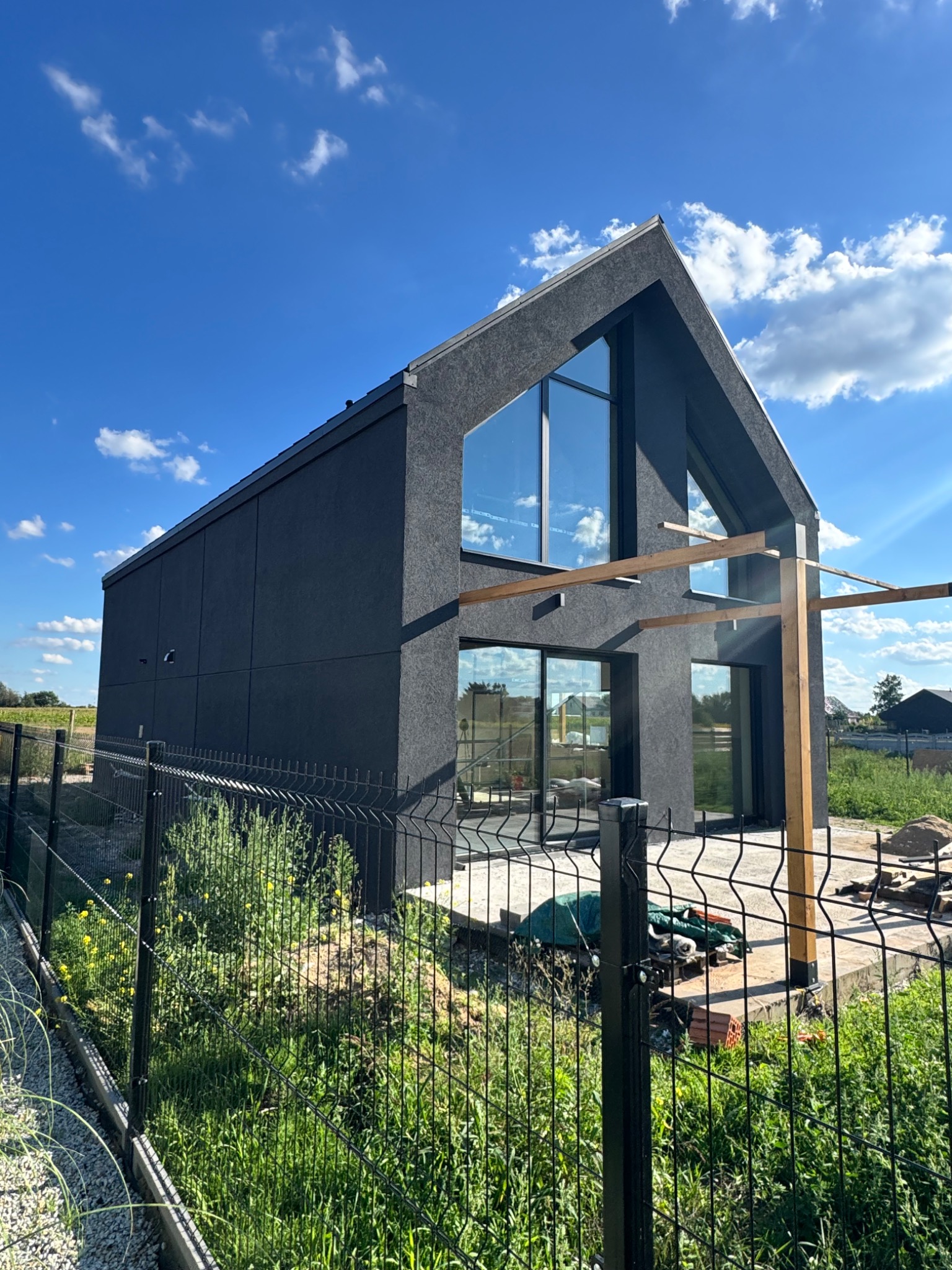 Nowoczesny dom z ciemną elewacją i dużymi oknami, widoczny zza metalowego ogrodzenia. Błękitne niebo z chmurami w tle. Elementy drewniane pergoli przy tarasie.