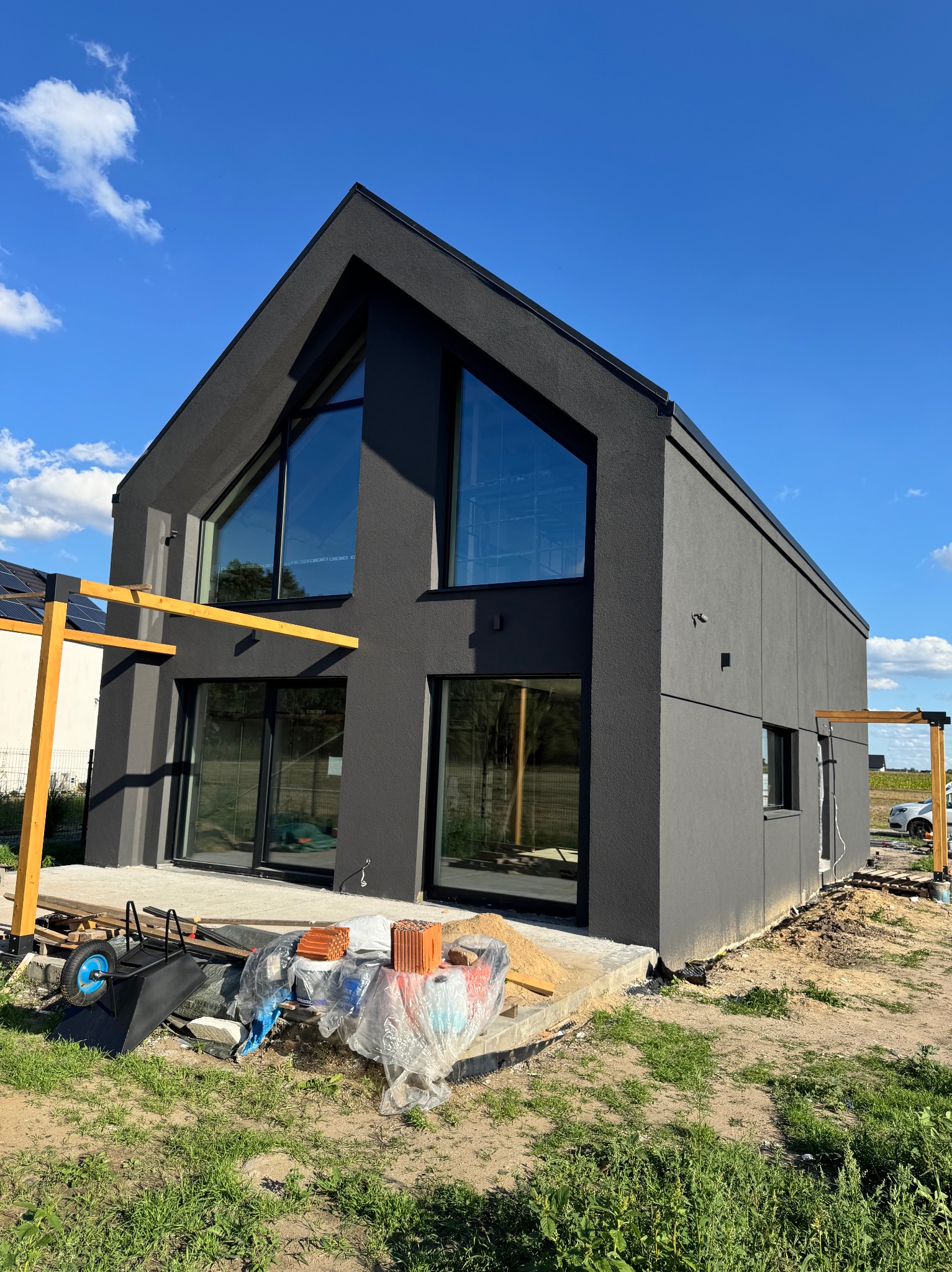 Nowoczesny dom z ciemną elewacją i dużymi oknami, w trakcie prac wykończeniowych. Widoczne materiały budowlane i elementy konstrukcji pergoli. Błękitne niebo w tle.