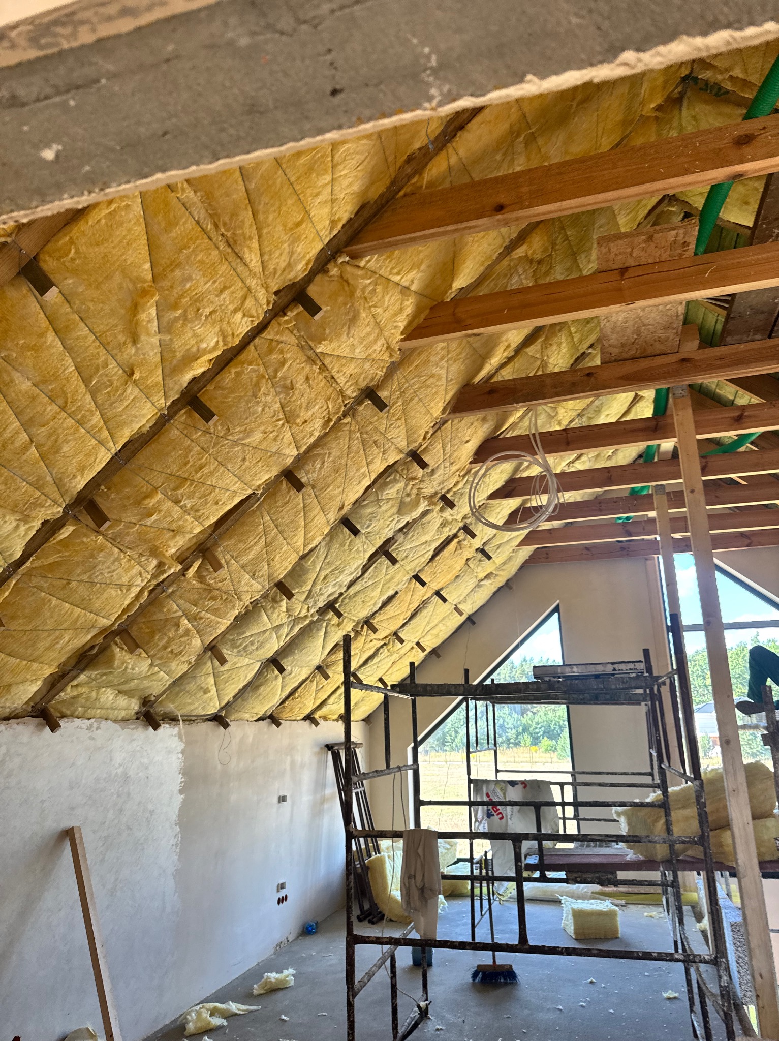 Skośny dach z drewnianą konstrukcją i żółtą izolacją, widok z rusztowania. Fragment otynkowanej ściany. Okno trójkątne w tle.