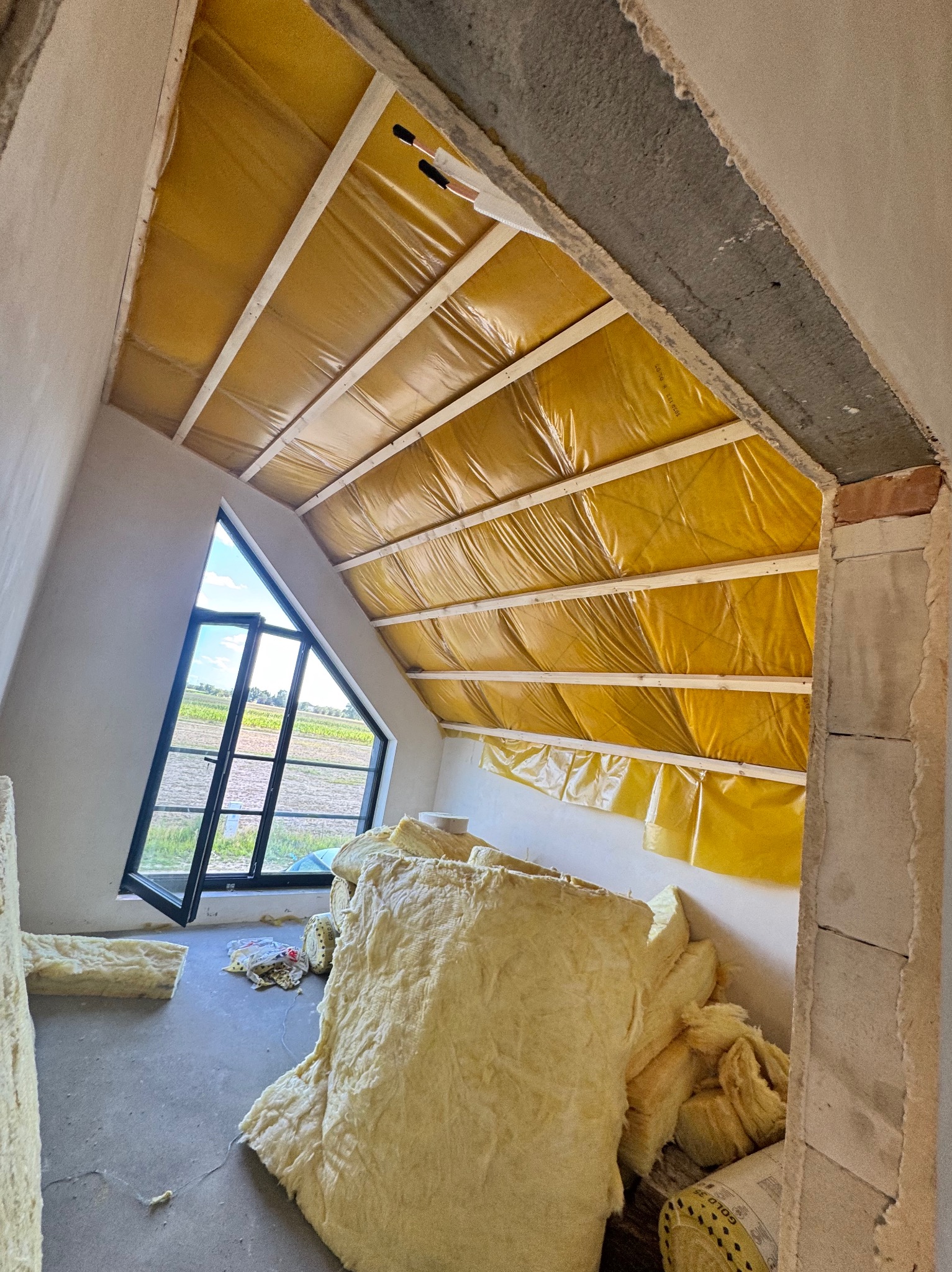 Poddasze w Pniewach w trakcie ocieplania. Widoczne belki, folia paroizolacyjna i wełna mineralna. Okno dachowe z widokiem na pole. Prace izolacyjne w toku.