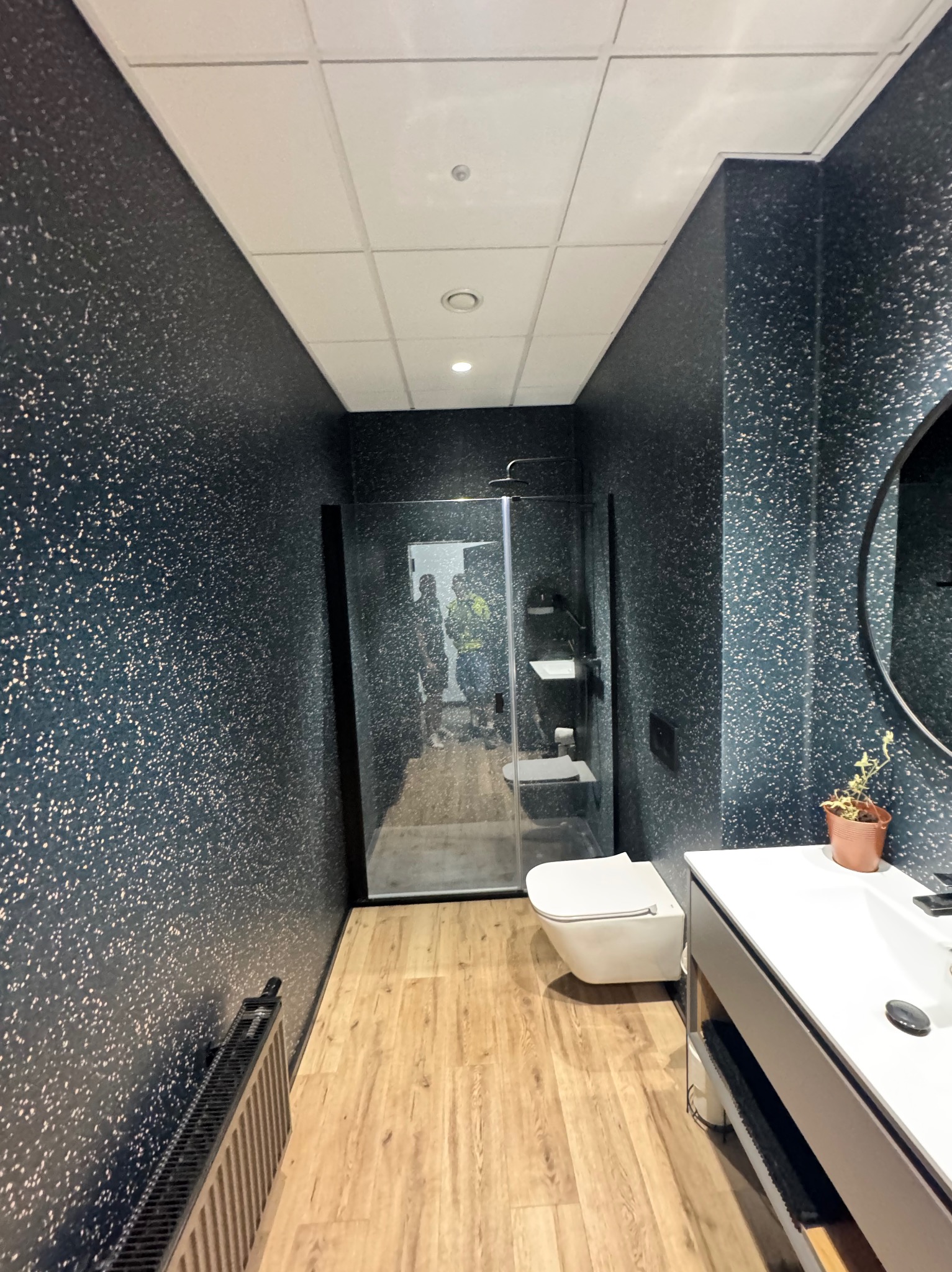 Nowoczesna łazienka z kabiną prysznicową typu walk-in, umywalką z szafką i toaletą. Ściany pokryte ciemną tapetą w jasne kropki, drewnopodobna podłoga. W tle widoczni pracownicy.