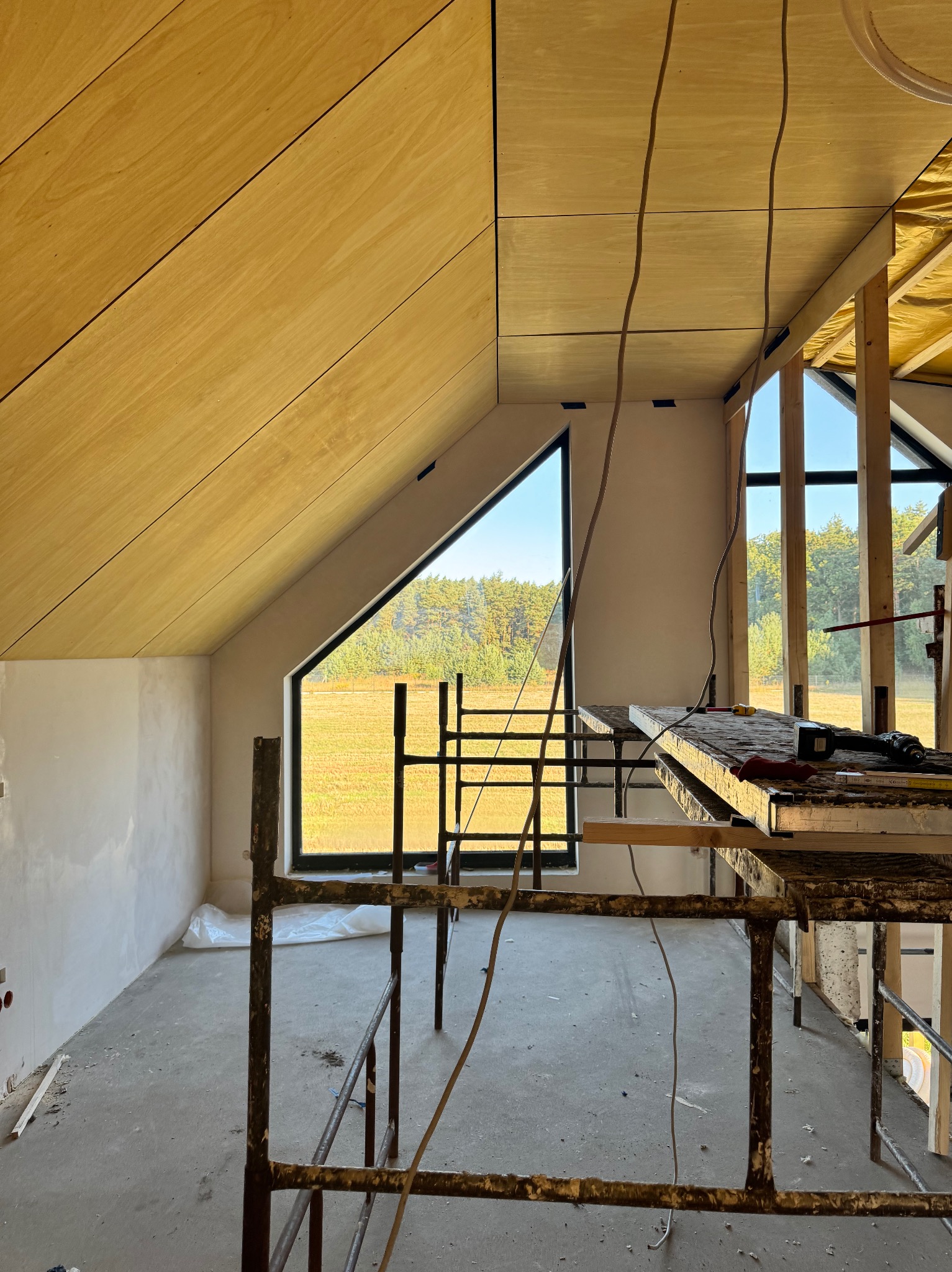 Poddasze w trakcie wykańczania: drewniana boazeria na skośnym suficie, trójkątne okno z widokiem na pole, rusztowania i narzędzia. Etap prac wykończeniowych.