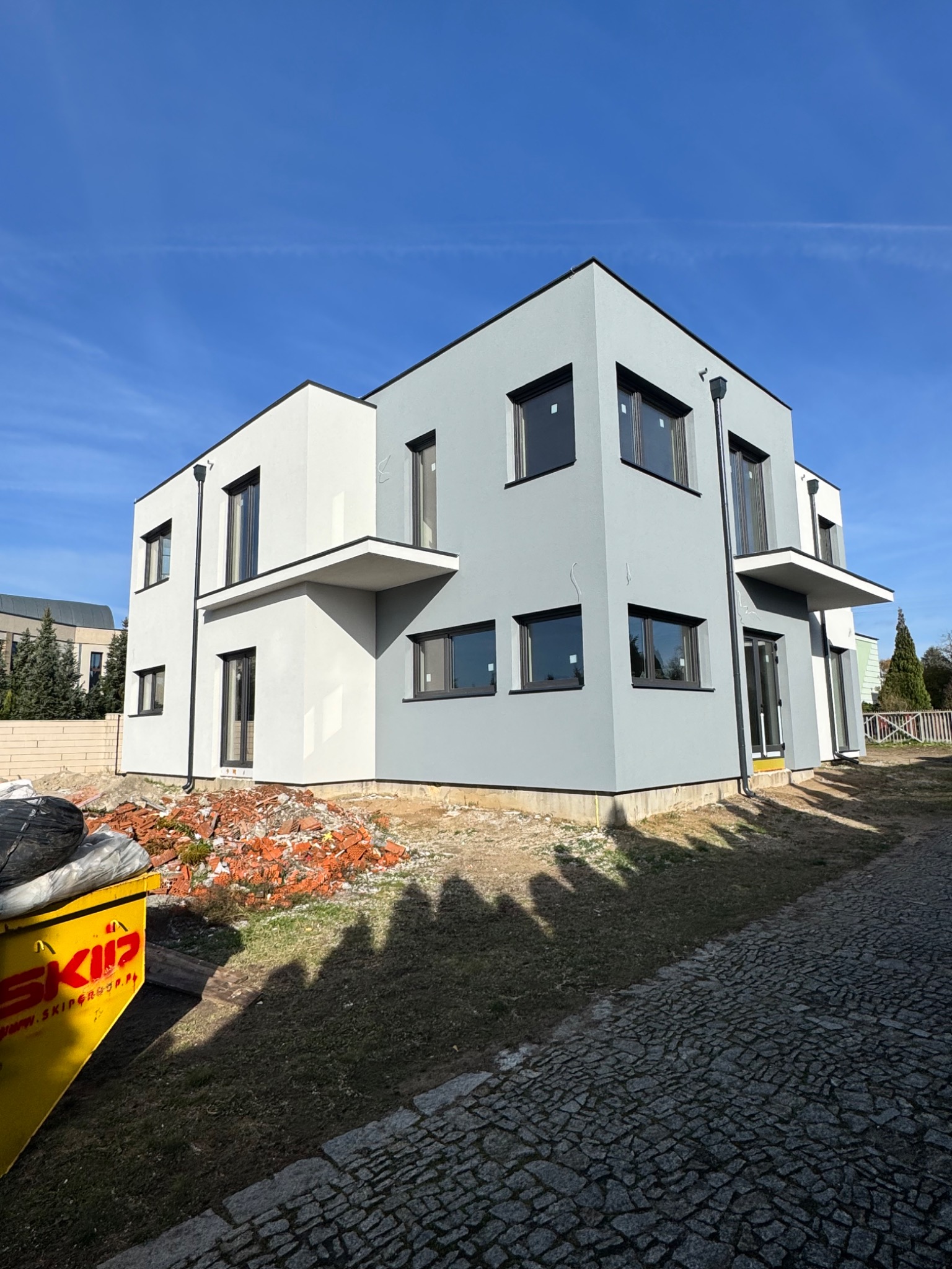 Dwukondygnacyjny dom z elewacją w odcieniach szarości i bieli, z czarnymi ramami okiennymi. Na pierwszym planie żółty kontener na gruz i kostka brukowa.