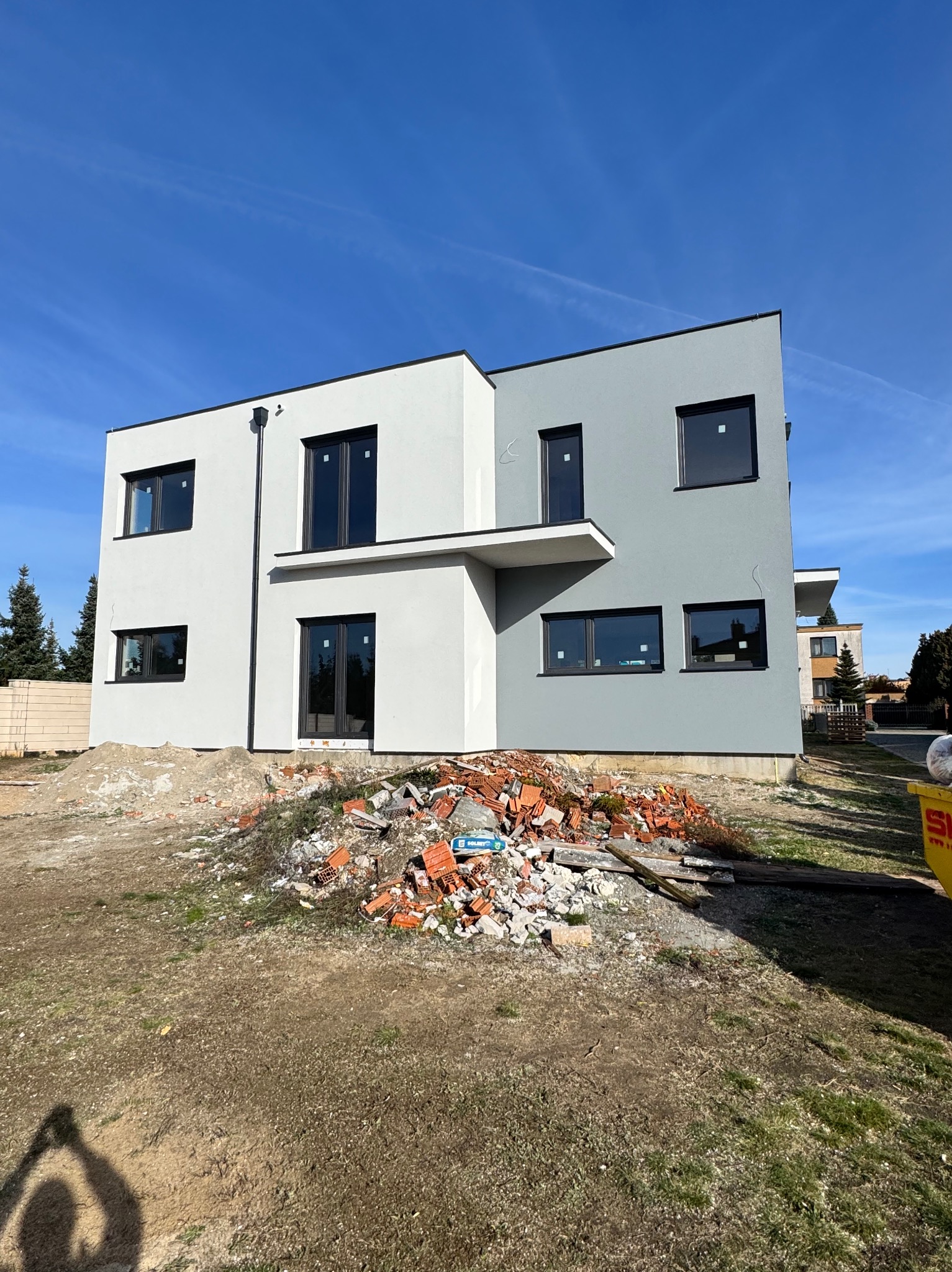 Dwukondygnacyjny dom z szarą i białą elewacją, czarnymi oknami i stertą gruzu przed budynkiem w słoneczny dzień. Minimalistyczny design.