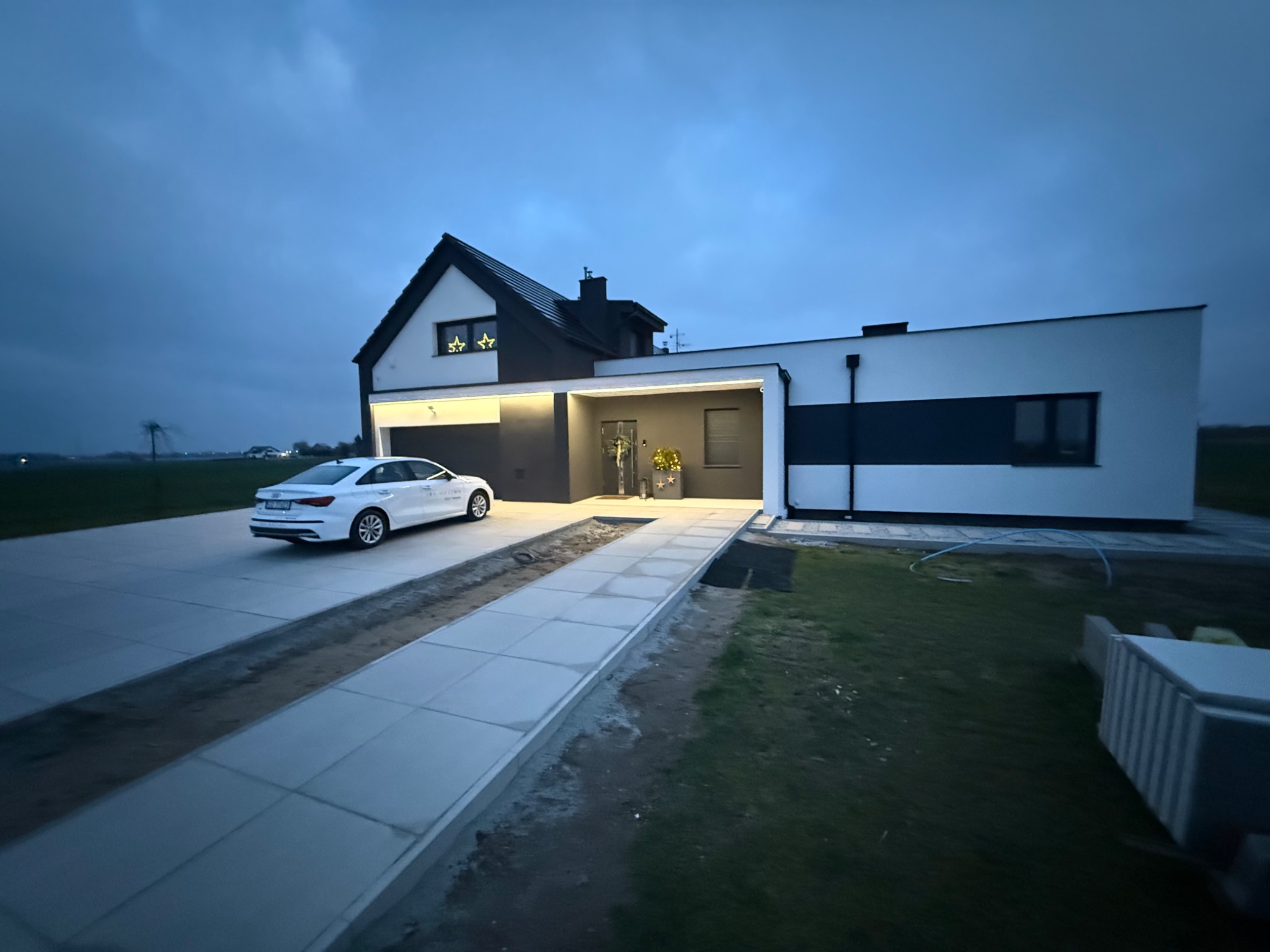 Nowoczesny dom jednorodzinny z białymi ścianami i czarnymi akcentami, oświetlony po zmierzchu, z podjazdem i zaparkowanym białym samochodem.