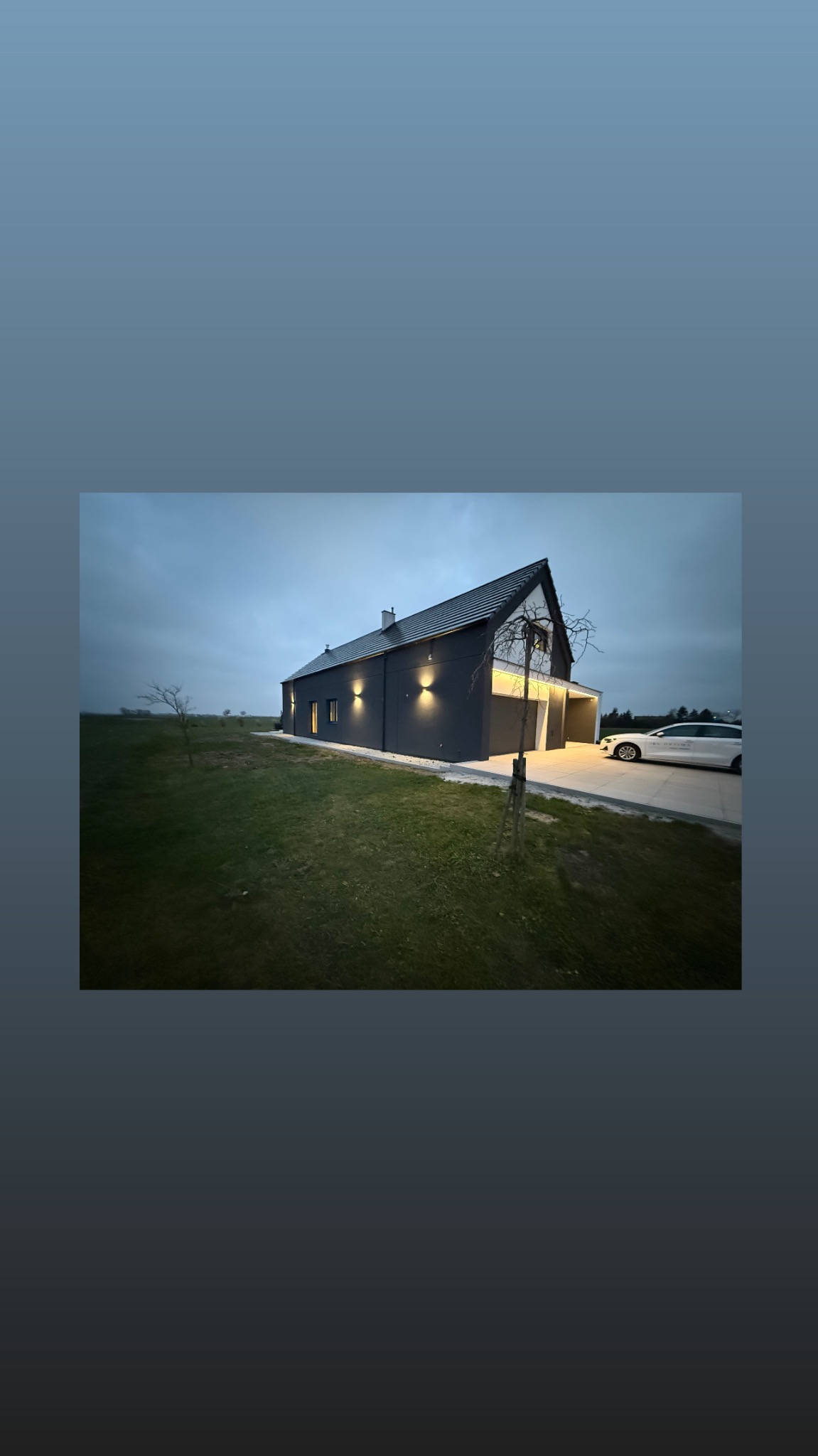 Nowoczesny dom jednorodzinny z szarą elewacją i oświetleniem zewnętrznym, widoczny z perspektywy ogrodu o zmierzchu. Na podjeździe stoi biały samochód.