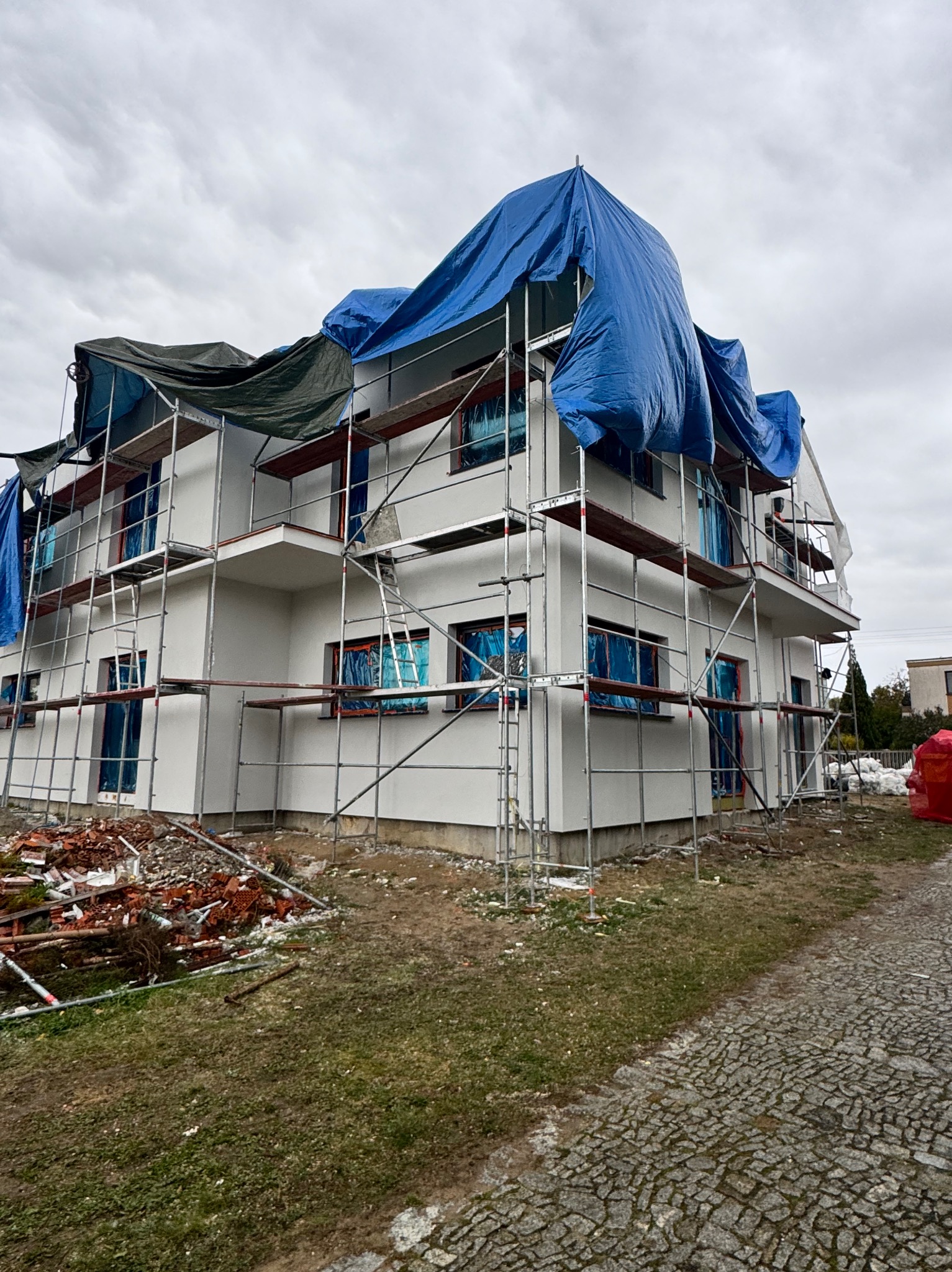 Biały dom w rusztowaniach, zabezpieczony plandekami, po świeżym malowaniu elewacji. Na pierwszym planie gruz i kostka brukowa, pochmurne niebo w tle.