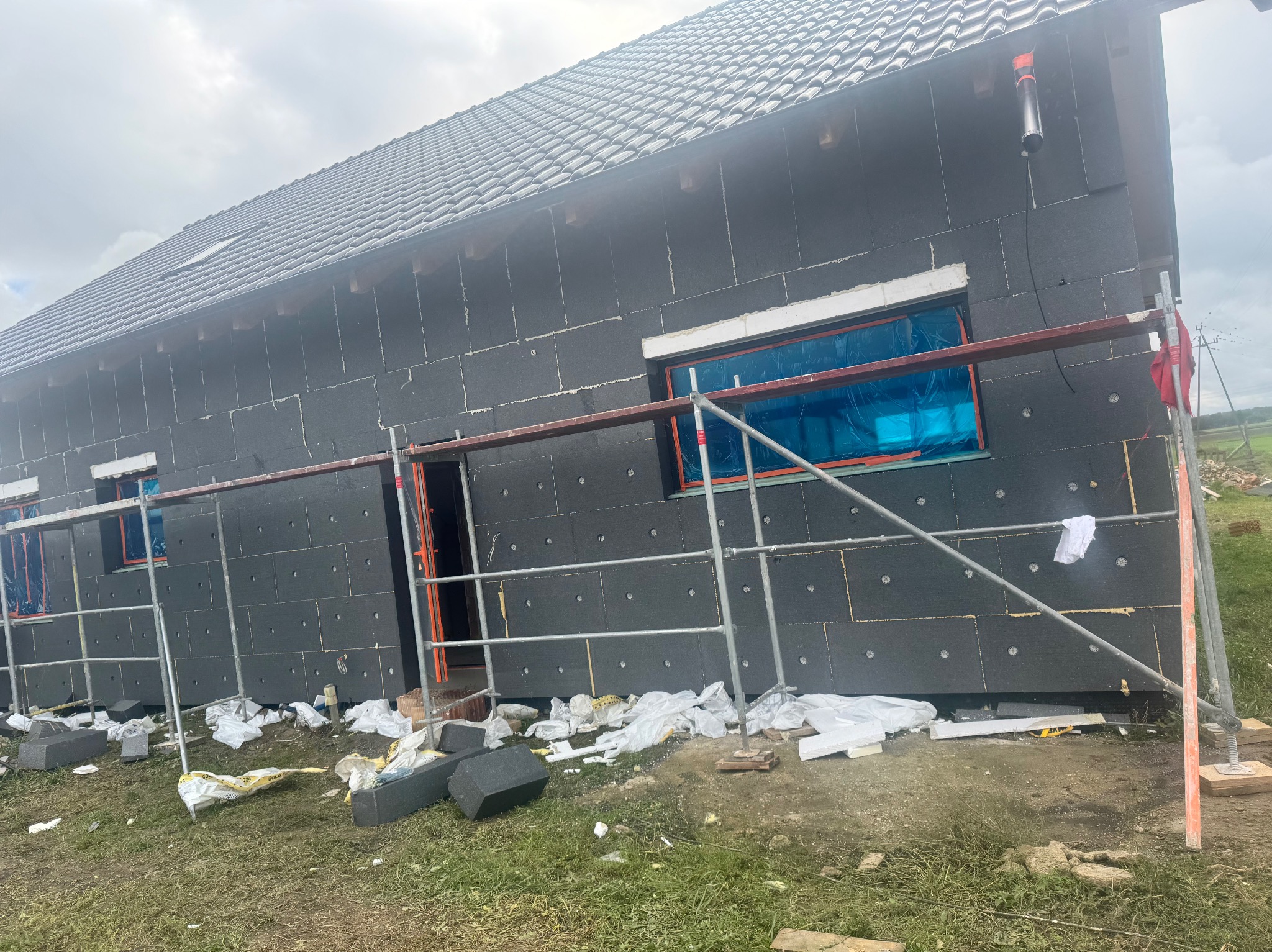 Dom w trakcie ocieplania elewacji czarnym styropianem, widoczne rusztowania i zabezpieczone okna folią. Na ziemi resztki materiałów budowlanych.