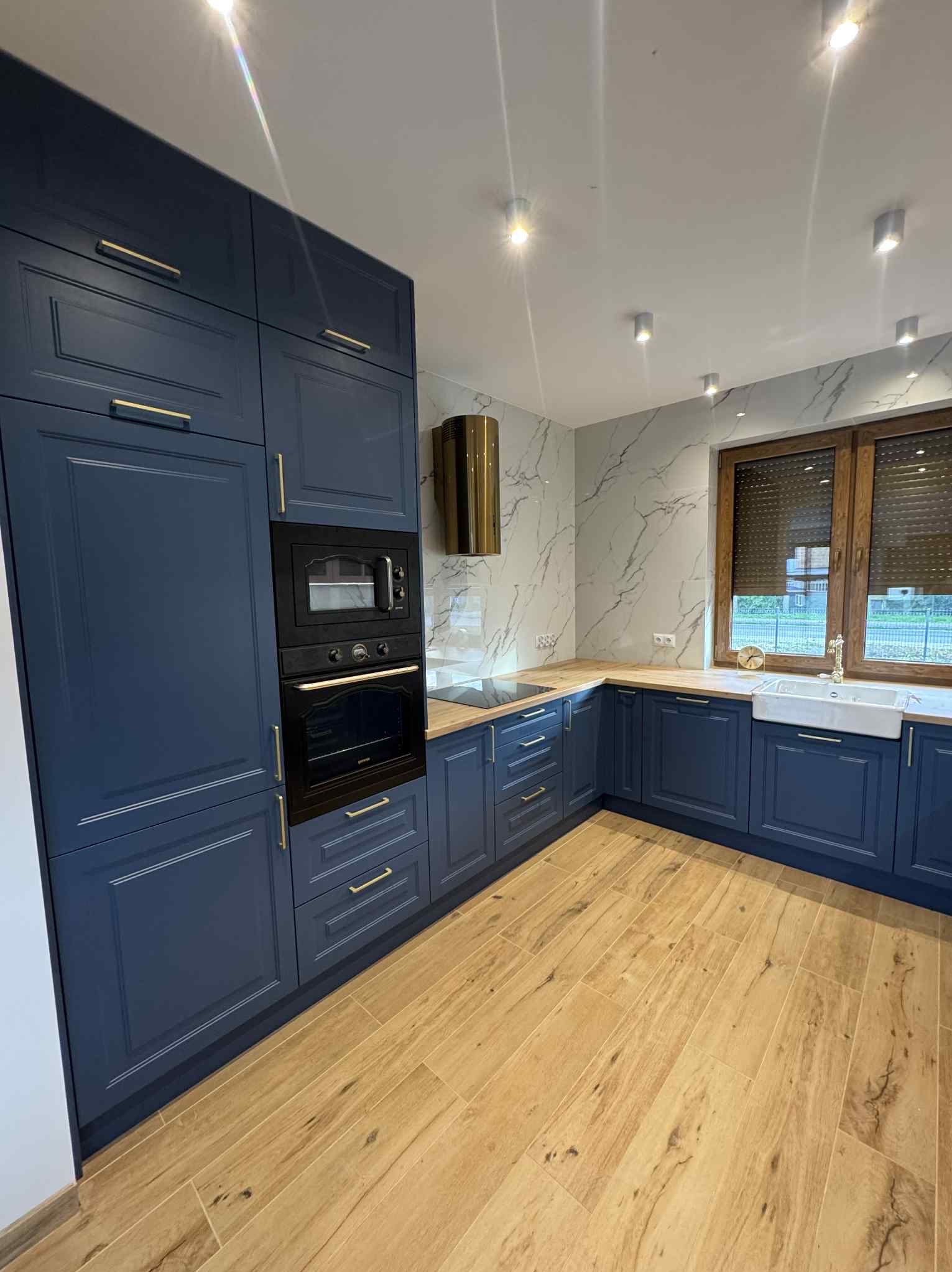 Granatowa zabudowa kuchenna z złotymi uchwytami, drewniany blat i podłoga imitująca drewno. Marmurowe płytki na ścianie i okno z drewnianymi roletami.
