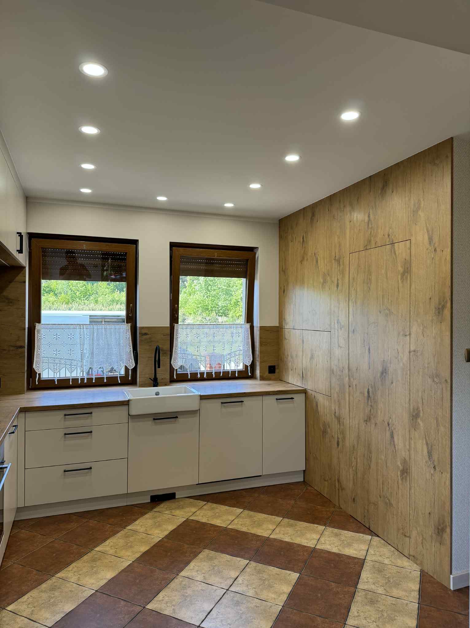 Jasna kuchnia z drewnianą ścianą i oknami z koronkowymi firankami. Uwagę przykuwają brązowo-beżowe płytki podłogowe i minimalistyczne, czarne uchwyty szafek.