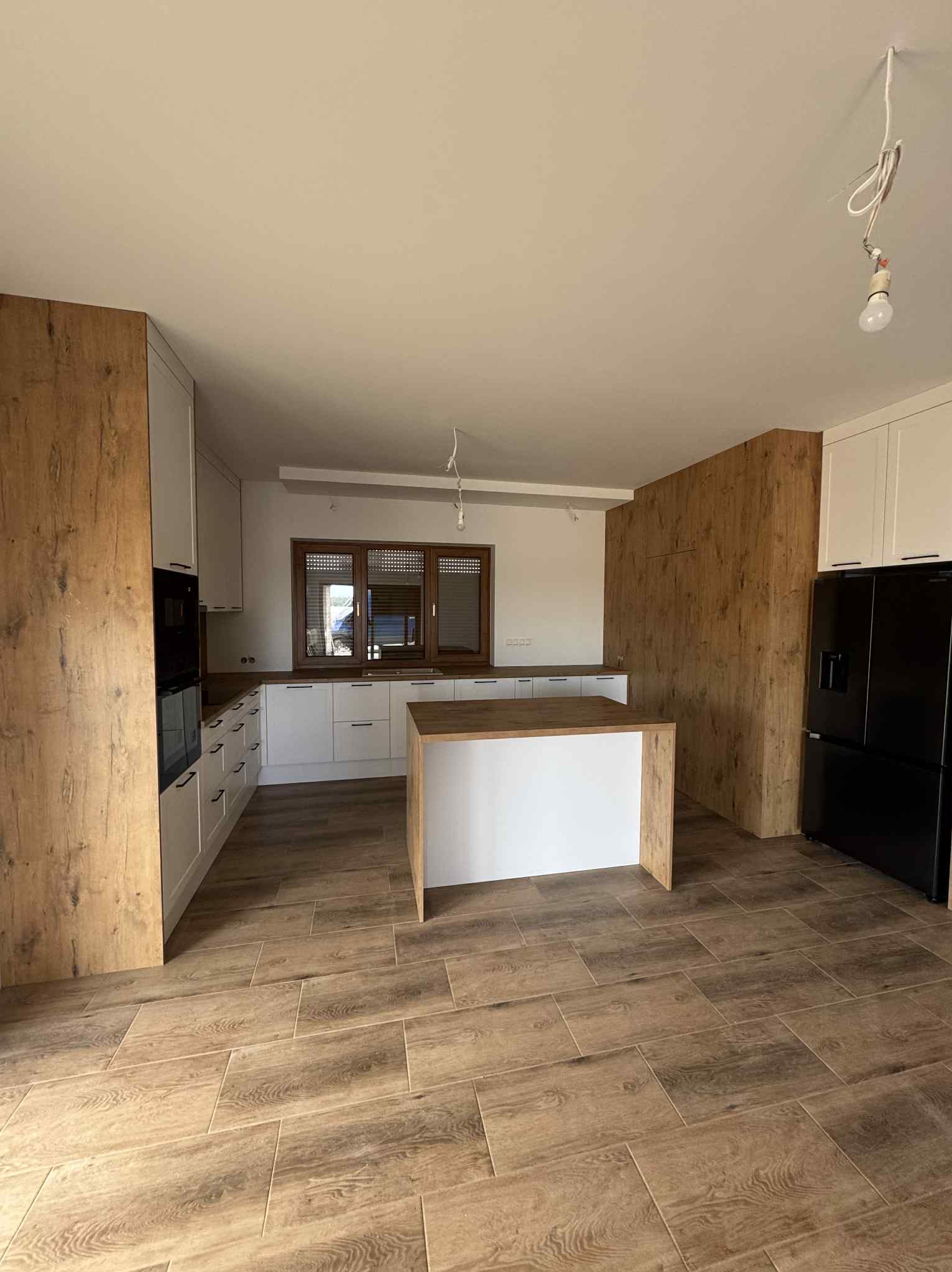 Jasna kuchnia z drewnianymi akcentami: wyspa, szafki i podłoga w odcieniach drewna. Białe fronty szafek i czarna lodówka. Widok na okno z żaluzjami.