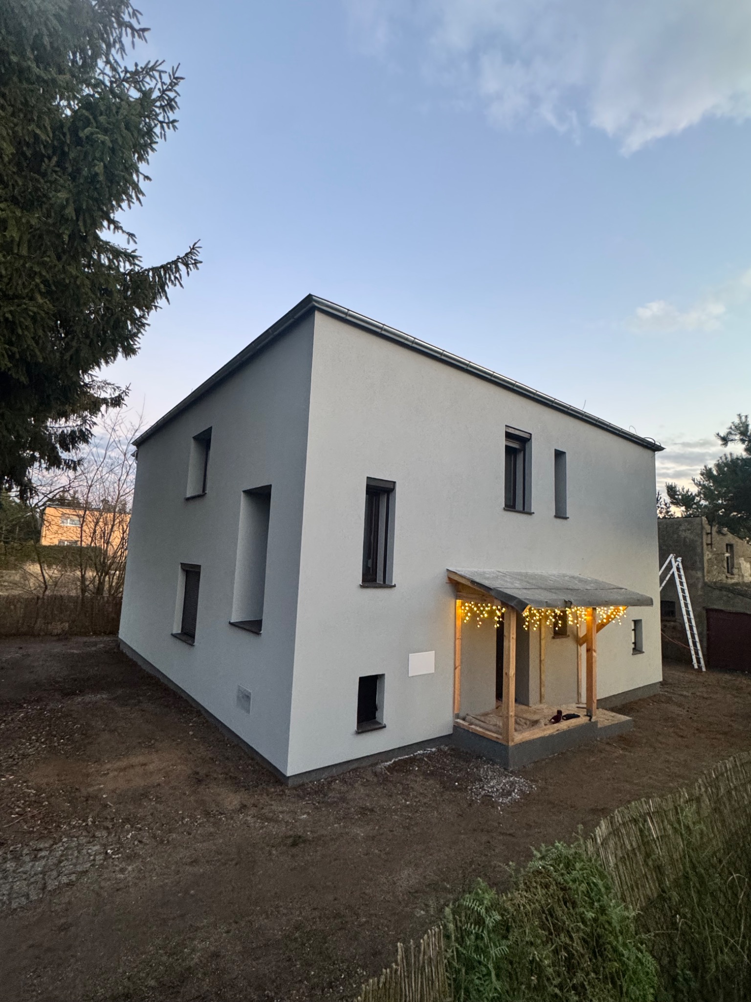 Nowoczesny, szary dom z płaskim dachem i drewnianym gankiem oświetlonym girlandą. Świeżo wykończona elewacja, surowy teren wokół. Ujęcie z dołu, w tle drzewa i drabina.