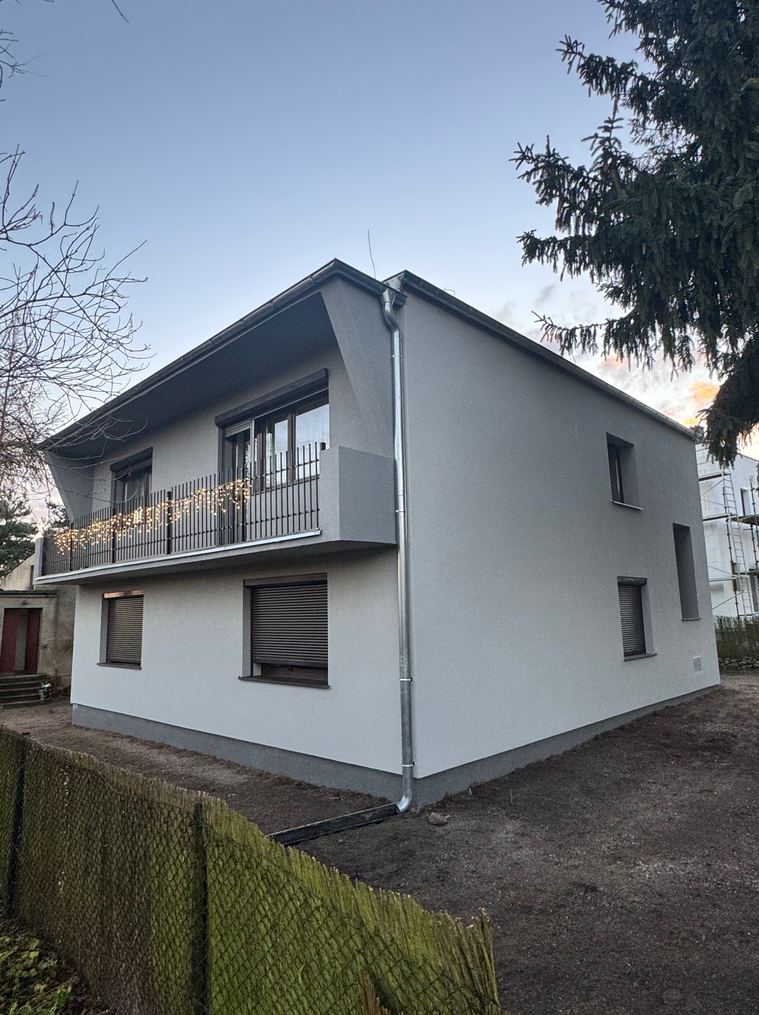 Dwupiętrowy dom z szarą elewacją i balkonem ozdobionym lampkami, widok z zewnątrz, po remoncie. Widoczne rynny i fragment ogrodzenia.