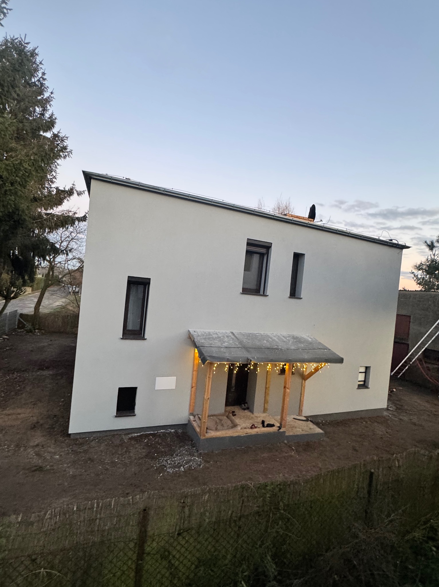 Nowoczesny, dwukondygnacyjny dom z jasną elewacją i drewnianym gankiem z lampkami, sfotografowany z lekkiej perspektywy, ukazujący świeżo zagospodarowany teren.