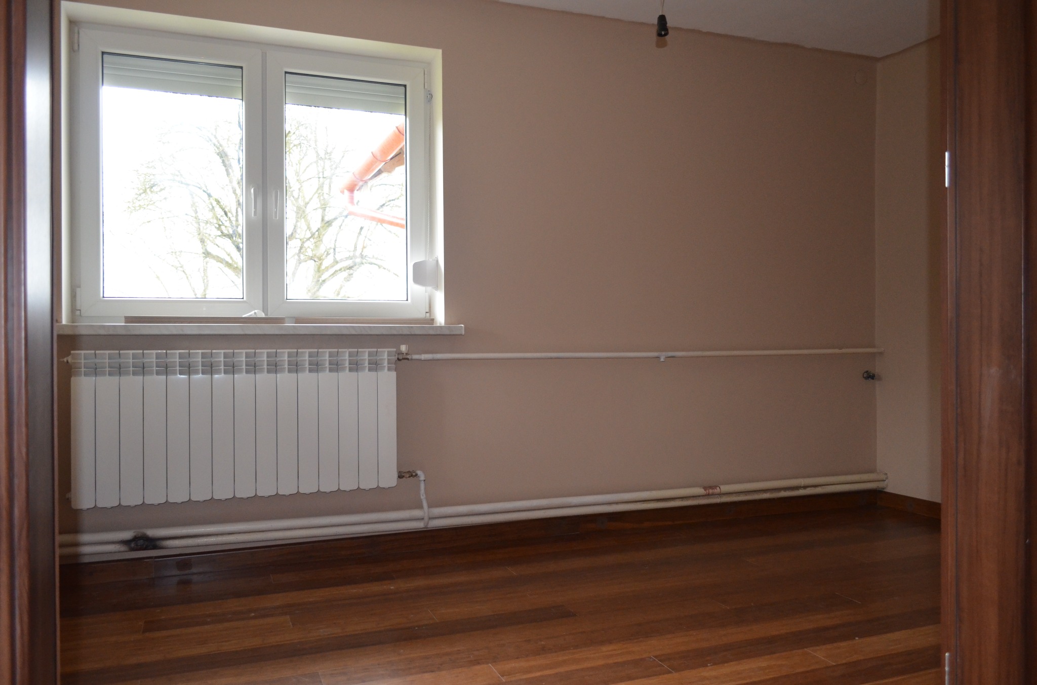 Pokój po remoncie: świeżo ułożona, ciemna podłoga z desek, beżowe ściany pomalowane do rogu, biały grzejnik pod oknem. Widoczne rury grzewcze wzdłuż ściany.