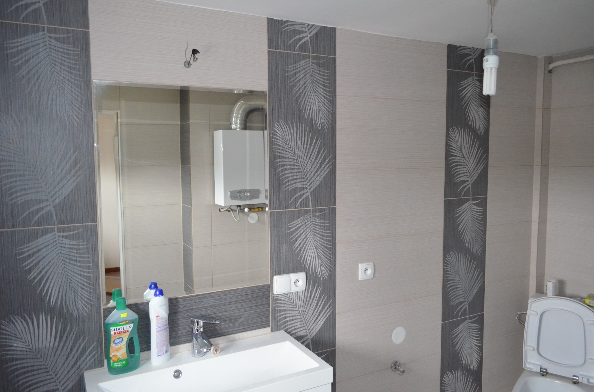 Jasna łazienka z szarymi akcentami: płytki z motywem liści palmowych ułożone pionowo, biała umywalka i toaleta. Widoczny bojler i niedokończone oświetlenie sufitowe.