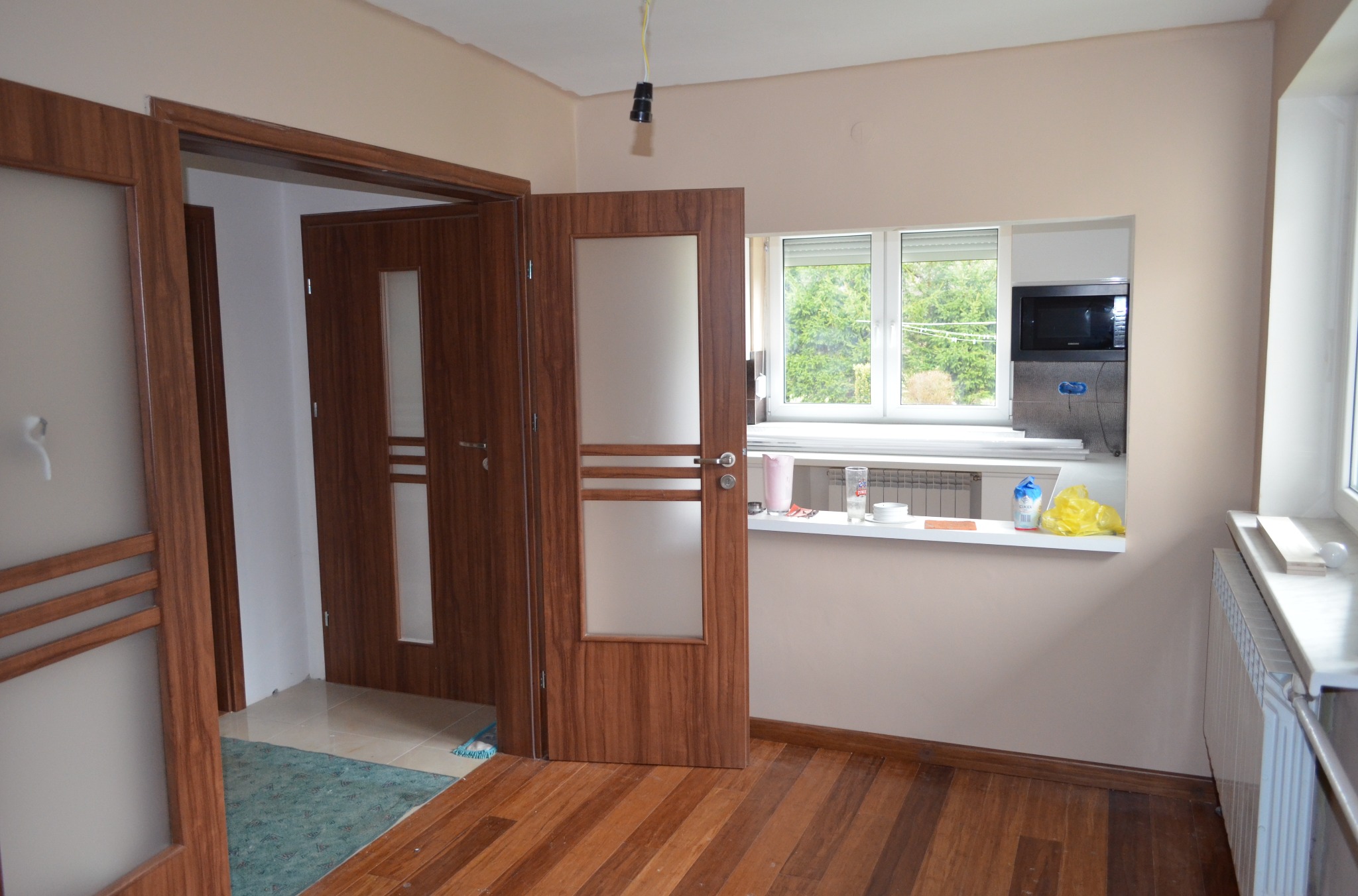 Wnętrze po remoncie: drewniana podłoga, drzwi z przeszkleniami, okno z widokiem na zieleń. Ściany w odcieniach beżu. Widoczny fragment instalacji elektrycznej.