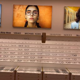 Mesa - Salon optyczny: ekspozycja okularów różnych marek (Miu Miu, Versace, Michael Kors, Swarovski, Gucci, MaxMara) na białych półkach, z reklamami wizualnymi powyżej.