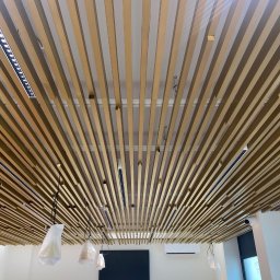 Mesa - Dekoracyjny sufit z drewnianych listew i zamontowanym oświetleniem LED. Lampy wiszące w folii ochronnej. Widoczny klimatyzator i kamera przemysłowa.