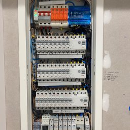 Mesa - Rozdzielnica elektryczna w białej obudowie, widoczne liczne bezpieczniki, przewody i oznaczenia. Ściana obok z notatkami ołówkiem.